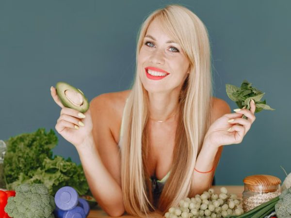 Blonde woman promotes healthy eating with fresh vegetables and fruits on display.